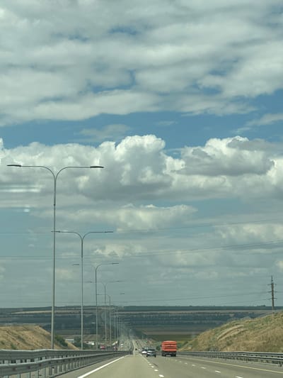 The image depicts a view from a highway in the vicinity of Grushevskaya, Russia, on a cloudy day. The highway stretches into the distance, with multiple lanes and a metal guardrail on the right side. Tall lampposts line the left side of the road. In the background, rolling hills covered with dry grass and scattered vegetation are visible, along with a band of trees and agricultural fields. Several vehicles, including cars and a large orange truck, are driving on the highway. Power lines are strung across the sky. The sky is filled with a mix of white and gray clouds.