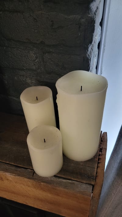 The image depicts three unlit, ivory-colored pillar candles of varying heights and widths, arranged on a rustic wooden surface. The candles are cylindrical with slightly softened edges at the top, and each has a visible black wick protruding from its center. They are positioned close together, with the largest candle on the right, a medium-sized one behind and to the left, and the smallest in the foreground to the left. The wooden surface appears weathered, with visible grain and some cracks. In the background, a dark, textured wall, possibly brick or stone, is present, illuminated by a light source from the right that casts a soft glow on the candles and the edge of the wooden surface. The overall lighting suggests an indoor setting, potentially a home or a decorative display. There is no indication of the city of Terrebonne, Canada, within the image.