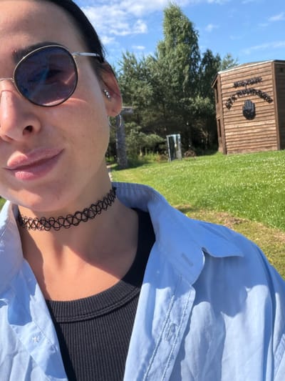 A woman with dark hair, wearing round sunglasses, a black choker, and a light blue unbuttoned shirt over a black tank top, is taking a selfie outdoors on a sunny day. She is looking at the camera and smiling. In the background, there is a grassy area with scattered white flowers, trees, and a wooden building with a sign. The scene appears to be in Rushinovo, Russia, possibly at an outdoor attraction or park.