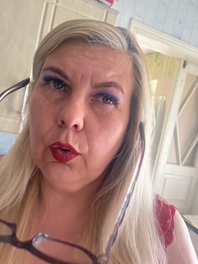 In the city of Pervomaisk, Ukraine, an image depicts a close-up of a woman's face. She has long, blonde hair and is wearing bright blue eyeshadow and red lipstick. Her mouth is slightly open, and she is holding a pair of glasses up to her face, obscuring parts of her eyes and nose. The background suggests an indoor setting, possibly a home, with indistinct furniture and a doorway visible. The lighting is bright, indicating daytime.