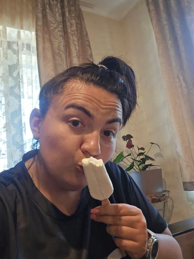 An individual with dark hair tied in a ponytail is captured in a close-up, indoor photograph taken in the city of Rudnik, Bulgaria. The person, viewed from the chest up, is seen holding and consuming a white popsicle from a wooden stick, with their lips positioned at the top of the frozen treat. Their right eye is clearly visible, looking directly at the camera, while their left eye is partially obscured. Wrinkles are evident on their forehead. They are wearing a dark-colored, short-sleeved v-neck shirt, and a silver-toned chain necklace is visible around their neck. A silver-colored smartwatch with a dark face is present on their left wrist. Their fingernails on the hand holding the popsicle are painted pink. The background features a window on the left with white patterned sheer curtains and heavier patterned light-colored drapes. Similar drapes are visible on the right side of the frame, along with a light-colored wall. A potted plant with dark green leaves and a red bloom in a grey rectangular pot is situated to the right of the person.
