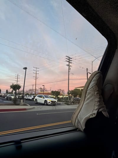 Inside a vehicle, a white sneaker rests atop the window frame, obscuring part of the view. Outside, utility poles line a road with parked cars, silhouetted against a pink and blue twilight sky. The photograph, seemingly taken from within the car, captures a casual, possibly irreverent, moment against the backdrop of a serene dusk.