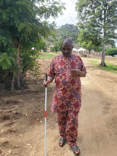 A man, who appears to be visually impaired, stands outdoors in Kabwir, Nigeria. He is wearing a traditional maroon and gold patterned garment and black sandals. He is holding a white cane with a red band near the handle, suggesting he is navigating his surroundings. The ground is a dusty, sandy path with scattered rocks and debris. To the left of the frame, a large tree with abundant green foliage dominates the scene. In the background, there are a few buildings, some appearing to be under construction, and a mountainous landscape. The sky is partially visible and appears to be overcast, suggesting a cloudy or possibly rainy day. The overall setting is rural and appears to be daytime.