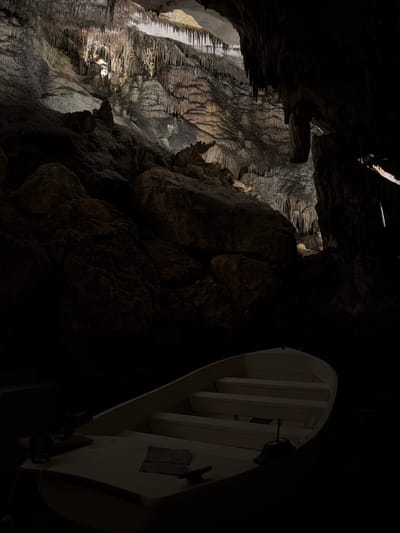 The image captures a dimly lit cave interior, possibly located in Manacor, Spain. The left side features a wall formation with stalactite-like structures, illuminated by a light source. A large rock formation occupies the center of the image. To the right, a white, framed object hangs from the cave ceiling, partially obscured by darkness. The environment is dark and cavernous. There are no visible people in the scene. The overall impression is that of a natural cave formation with an added, man-made structure.