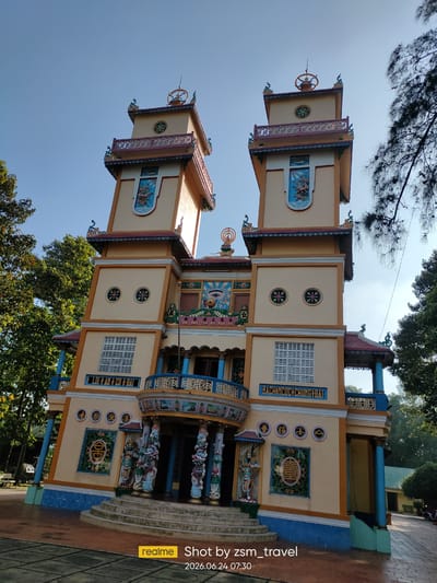 This is a medium-shot, eye-level photograph of a large, ornate temple in Gò Dầu, Vietnam. The temple is painted a vibrant yellow and adorned with blue and red accents. It features two prominent towers with decorative spires and colorful murals. Intricate carvings and statues are visible at the base of the temple, surrounding a set of stairs leading to the entrance. The architecture suggests a Buddhist or Taoist influence, with symbols and figures commonly found in these religions.
The image is taken during the day, under a clear blue sky, indicating fair weather. Sunlight illuminates the temple, casting subtle shadows and highlighting its detailed design. Lush green trees frame the sides of the temple, adding a natural element to the scene. The overall mood is serene and majestic, conveying a sense of peace and spiritual significance.
Visible text within the image includes inscriptions in Vietnamese on the temple walls, and a watermark at the bottom right corner displaying "realme Shot by zsm_travel 2026.06.24 07:30". The text on the temple appears to be religious or directional phrases.