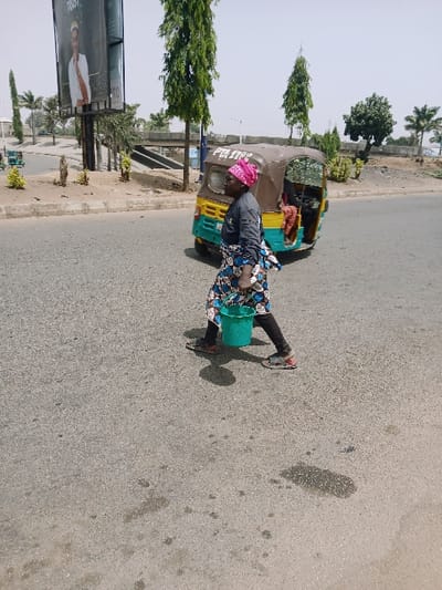 An adult female walks across an asphalt road in Kadoma, Zimbabwe, from left to right. She is dressed in a pink patterned headwrap, a dark long-sleeved top, dark trousers, and a blue and white patterned skirt, carrying a bright green plastic bucket in her left hand. Her footwear consists of open-toed sandals. Behind her, a stationary brown, yellow, and green three-wheeled auto-rickshaw, bearing the text 'PILOT STAR,' is visible. The background features a large billboard displaying a man in a white garment, tall green trees, an elevated road structure, and a dirt median. The road surface is asphalt with visible wear and stains, under a clear sky.