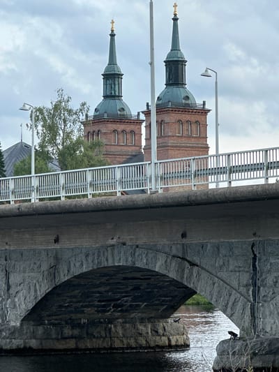 The image shows the twin spires of the Vammala Church in Vammala, Finland, as viewed from beneath a stone arch bridge. The church spires, topped with golden crosses, are made of red brick and have green copper roofs. A white metal fence lines the top of the bridge, which is constructed of large grey stones. A body of water flows under the bridge, reflecting the overcast sky. Several streetlights are visible on the bridge. The lower part of the image shows the underside of the bridge with its stone arch and the water below.
