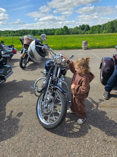 A young child wearing a brown hooded sweatshirt and pants is standing next to a motorcycle, seemingly interacting with the handlebars or controls. The motorcycle is chrome and has a blue gas tank. The scene appears to be in an open area with other motorcycles parked nearby. A field with yellow flowers and trees is visible in the background, under a sky with scattered clouds. The exact location within Borodinskogo Muzeya, Russia, is difficult to determine from the provided image alone, but it is likely an outdoor area near parking for visitors.