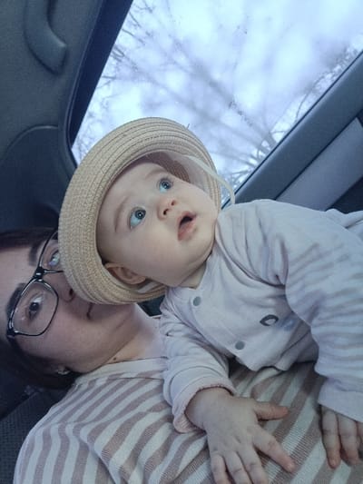This image depicts an adult and an infant inside a vehicle in Simakovo, Russia. An adult, visible from the chest up on the left, wears dark-rimmed eyeglasses with black frames and a light beige and white striped long-sleeved shirt. An infant, wearing a light beige straw hat and a pale pink long-sleeved outfit with gray snap buttons down the front and a small dark oval design on the chest, is positioned on the adult's torso. The infant has light blue eyes, an open mouth, and is looking upward towards the upper frame. The adult also appears to be looking in a similar upward direction. Through the vehicle's windshield or window, a cloudy sky and blurred, bare tree branches are visible. Dark gray vehicle interior trim is present along the upper left and right edges of the frame.