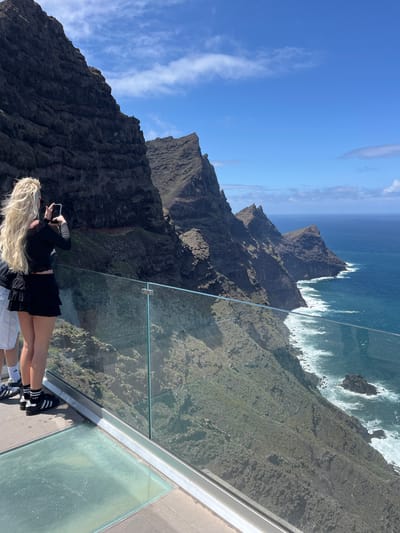 The image depicts a scenic overlook at the Mirador del Balcón in La Aldea de San Nicolás, Spain. Two people are visible on the glass-floored platform. One person, with long blonde hair, is lying down and taking a photo with a smartphone. They are wearing a black long-sleeved top, a black skirt, black and white striped socks, and black Adidas sneakers. The other person is partially visible, wearing white shorts and Adidas sneakers. The overlook is positioned on a cliffside, offering a panoramic view of rugged coastal terrain, cliffs plunging into the Atlantic Ocean, and crashing waves. The sky is clear blue with wispy clouds.