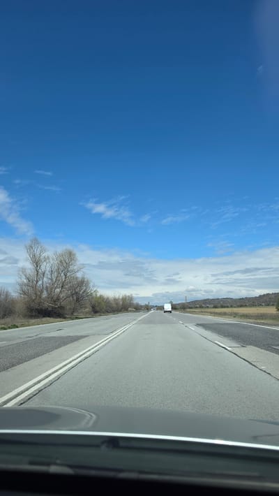 A view from inside a vehicle, traveling on a straight, two-lane road. The road is asphalt with double white lines dividing the lanes. There is a white truck visible in the distance, traveling in the same direction. Vegetation is visible on both sides of the road, with bare trees. The sky is a clear blue with scattered white clouds. The location of the photograph cannot be determined from the image alone.