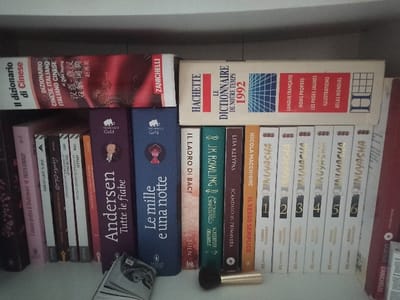 This image displays a section of a bookshelf in an indoor residential environment within San Marzano sul Sarno, Italy. The shelf is light-colored, likely white, presenting two levels of organized books.
On the upper shelf, two large dictionaries are visible: a "Dizionario di Cinese" (Chinese Dictionary) by Zanichelli with a red and white spine on the left, and a "Hachette Le Dictionnaire de Notre Temps 1992" featuring a white spine with a blue grid pattern on the right.
The lower, main shelf contains a diverse collection of books arranged vertically. From left to right, visible spines include works by Shakespeare, Felicity Kingsley, Spencer Lewis, and Agatha Christie. Centrally located are two prominent, dark-bound books from the "Mammut Gold" series: "Andersen Tutte le fiabe" (Andersen All the Fairy Tales) in dark purple, and "Le mille e una notte" (One Thousand and One Nights) in dark blue. Following these are an orange book titled "IL LADRO DI BACI" by SILEN, a green-spined book by J.K. Rowling (with "Grindelwald" visible), a dark red book by Lisa Kleypas titled "SCANDAL IN SPRING", and an orange book by Nicola Macchione titled "IL SESSO SEMPLICE". The rightmost section features a sequence of six white-spined manga volumes, numbered 1 through 6, all titled "INUYASHA" by Rumiko Takahashi. A tall, thin red book by Zanichelli is partially visible on the far right.
In the foreground of the lower shelf, a small, partially open item, possibly a booklet or folded papers, displays a black and white image of a person's face, resembling a man with a mustache. Adjacent to it on the right, a makeup brush with a black handle and a gold ferrule lies horizontally across the shelf.