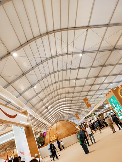 The image captures the interior of a large, arched exhibition tent or hall in Al Machouar – Stinia, Morocco. The expansive ceiling is made of white fabric panels supported by a visible metallic grid structure with crisscrossing tension cables, illuminated by numerous recessed circular lights.
The scene is bustling with a diverse crowd of people, casually dressed, walking and interacting throughout the hall. Various exhibition booths line the spacious light-colored floor. On the left, a tall, modern-looking booth features a white panel with a prominent curved red design and partially visible text, including "DANS". A large, white, flexible duct runs high along the left side of the tent structure.
Towards the center, a large, dome-shaped booth with a wooden-like finish displays illuminated Arabic text. On the right, a striking booth with a tall, rectangular display panel shows green and white graphics and Arabic script, with "Azur" partially discernible. Adjacent to it is a brown structure resembling castle battlements. Orange banners with Arabic text and "7 B" are suspended from the ceiling.
The overall impression is of a lively public event, such as a market, fair, or exhibition, taking place indoors under bright artificial lighting. The presence of Arabic script on signage reinforces the location context in Morocco.