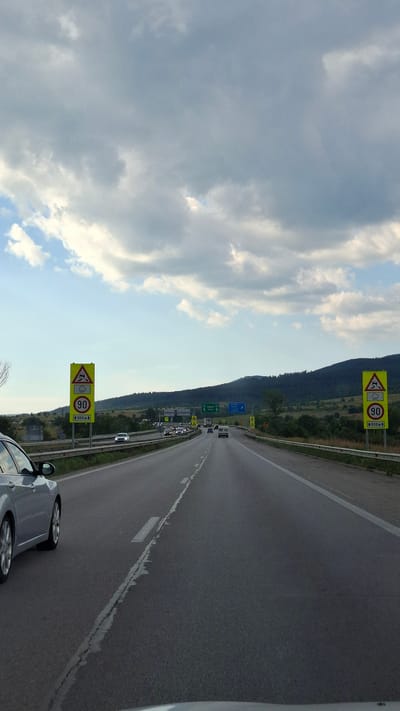 The image shows a view from a vehicle driving on a highway. The highway has multiple lanes and a barrier on the right side. There are speed limit signs and warning signs along the road. In the distance, there are hills and trees. The sky is cloudy. The car in the foreground on the left is silver. There are other cars on the highway, some in the distance and some closer. The scene is consistent with a highway outside of a city, likely in a rural or semi-rural area. The location is specified as Mirovo, Bulgaria.
