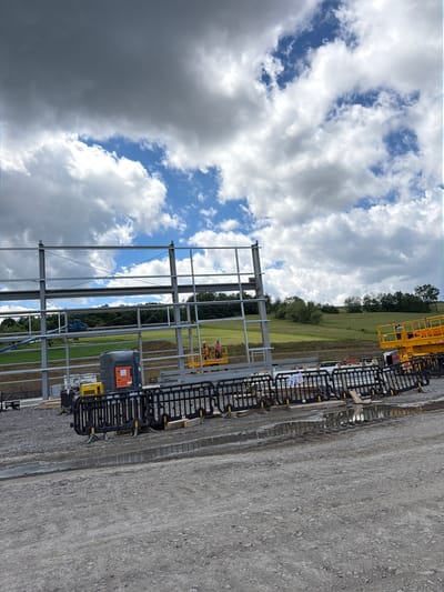 The image captures a construction site with a large steel structure in the foreground, set against a backdrop of a cloudy sky and a grassy hillside. On the right side of the frame, a yellow aerial work platform is visible. In the middle ground, a black and orange generator unit is situated next to a similar aerial work platform with two individuals wearing hard hats and safety vests. Black plastic barriers with yellow accents are placed in front of the generator and platform. Further back, a blue boom lift is parked on a grassy area, and a treeline borders the top of the hill. The ground in the foreground is covered in gravel, with a puddle of water visible. The location is identified as the city of Jodłownik, Poland.