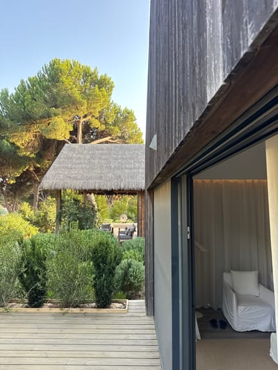 The image depicts an outdoor scene with a contemporary wooden building on the right, featuring a sliding glass door that reveals a sparsely furnished interior with white curtains and a white armchair. The building's exterior is clad in vertical wooden planks, and a wooden deck extends from the foreground. To the left of the building, lush green vegetation and mature pine trees are visible. In the mid-ground, a thatched-roof structure, resembling an open-air pavilion, is situated among the greenery, with outdoor furniture visible underneath. The sky above is clear and blue. This scene is located in Grândola, Portugal.