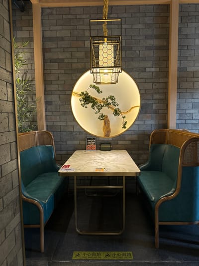 The image displays an interior dining or lounge area, likely within a restaurant or cafe in Fangshan District, China. The setting features a contemporary design with traditional Chinese aesthetic influences.

The walls are constructed from grey rectangular bricks, and a light wooden beam horizontally spans the mid-ground. Below this beam, artificial green foliage resembling bamboo leaves adds a natural element.

A prominent circular backlit art piece on the back wall showcases a gnarled, bonsai-like tree with green leaves, seemingly growing from a rocky landscape, emitting a warm, soft glow. To its left, a modern geometric lantern with a golden frame and hexagonal patterns hangs from the ceiling, providing additional ambient lighting.

In the foreground, two comfortable teal-colored upholstered armchairs, or booth seats, with wooden frames and rattan detailing on their backrests, are arranged opposite each other. Between them is a rectangular table with a light-colored, possibly marble-patterned, top and a dark metal base. Small stickers or papers, likely menus or promotional items, are visible on the table surface. A small rectangular sign with pictorial representations of food items is also visible on the brick wall above the table.

The floor is a dark, textured surface. On the far right, a yellow warning sign is visible, displaying the Chinese characters "小心台阶" and the English phrase "Caution Watch your step," along with a pictogram of a person falling, indicating a step or change in floor level.

No people are present, suggesting a quiet or unoccupied moment. The overall atmosphere is calm and inviting, with a focus on thoughtful design and lighting. The indoor setting does not provide weather details, but the lighting suggests a controlled environment, potentially an evening ambiance.