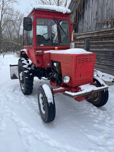 A weathered red tractor, consistent with a Soviet-era model, is stationary in a snow-covered rural setting within Карабоўскі сельскі Савет, Belarus. The tractor's cab roof, hood, and tires are dusted with fresh snow. It features a full cab, a single amber rotating beacon on the right side of the hood, and two round headlights. A snow plow blade is mounted to the left side of the chassis, positioned in front of the rear wheel. The exhaust pipe extends vertically on the right side of the engine compartment. Directly behind the tractor, to its right, is a rustic wooden structure with vertically oriented planks and a snow-dusted roof. The wider background on the left consists of leafless trees and brush under an overcast sky. The entire visible ground surface is uniformly blanketed in snow. No individuals are depicted.