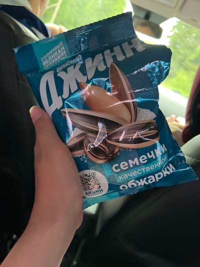 A person is holding a bag of "Обжарки" sunflower seeds in their hand, likely inside a vehicle. The bag is blue and features an image of sunflower seeds on the front. The text on the bag is in Russian, indicating that the product is intended for a Russian-speaking audience. The person's hand is visible and appears to be resting on their lap, likely inside a car. The background suggests that the location is outdoors, possibly near foliage visible through a car window. There is no indication from the image that the location is in Большие Кусты, Russia.