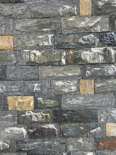 The image displays a close-up, full-frame view of a wall constructed from stacked, irregularly shaped stone tiles. The tiles vary in color, with dominant shades of gray, black, and some lighter, yellowish-brown accents. The texture of the stones is rough and natural, with visible grain and layered striations. The arrangement of the tiles is staggered, with vertical placement being the most prominent orientation, creating a visually textured surface. Sunlight appears to be hitting the wall from the side, casting shadows that highlight the uneven surfaces and depth of the stones. The lighting suggests it is daytime, likely outdoors given the natural stone material. No people, objects, or text are visible in the image. The setting is a stone wall, potentially as an exterior facade or an interior decorative element. The context provided indicates the location is Kadewatta, Sri Lanka.