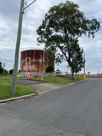 The image displays an outdoor scene, likely an industrial or semi-industrial area in Hamilton, Australia, under a lightly clouded sky during daytime. A large, mature tree with dense green foliage stands prominently on the left side of the frame. Behind and partially obscured by the tree is a massive, cylindrical storage tank, heavily rusted with a deep reddish-brown patina.

Large white text is clearly visible on the tank, reading "OMEGA CHEMICALS," with "SULPHURIC ACID" and "BOILER FUEL" inscribed beneath it. The lower section of the tank and an adjacent low structure are covered in various, illegible graffiti tags. Near the tank, two red circular signs with white '00's are also visible.

To the right of the tree and tank, a wide asphalt road extends into the distance. It features a yellow dashed center line and a white solid line on its edge. A concrete sidewalk runs parallel to the road, separated by a lush green grassy verge. This verge also extends along the base of the storage tank. In the immediate foreground on the left, a wooden utility pole rises. Further down the road, in the background, more industrial elements are visible, including what appear to be pink structures and a construction crane, indicating ongoing activity. Orange and white safety barriers are scattered along the verges and near the tank. No people are visible, and the scene is static, showcasing infrastructure and the natural environment.