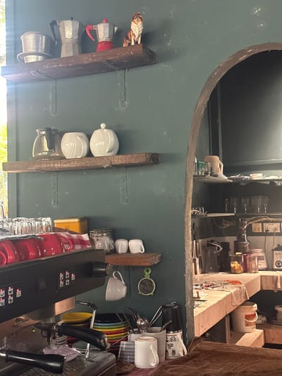 A cluttered workspace features a variety of items including coffee makers, cups, and an espresso machine. Coffee pots are nestled on a makeshift wooden shelf with a small dog figurine. Above is a dark recess with various machines and equipment. Other shelves hold sugar bowls and jars. A stack of mugs and plates sits near a black espresso machine, surrounded by utensils and miscellaneous items. The overall impression is one of a functional but disorganized coffee-making area.