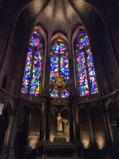 This is a wide-angle, low-light, interior shot of a cathedral, specifically the church of Saint-Germain in Urvillers, France. The focal point is a large, ornate stained-glass window filling the upper portion of the frame. The window is composed of numerous small, irregularly shaped panes of glass in vibrant colors, including red, blue, yellow, and green, set against a darker background. Below the window, a statue of a robed figure, possibly a saint or historical person, stands on a pedestal. The statue is illuminated by spotlights from the floor, casting shadows. The surrounding architecture features dark stone columns, arches, and vaulted ceilings, characteristic of Gothic architecture. The overall impression is one of grandeur and historical significance, with the colorful stained glass providing a stark contrast to the dark stone.