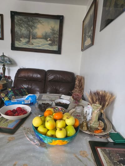 This interior image, set in Cwmbrân, United Kingdom, depicts a warm and decorated living room or sitting area. The scene is dominated by a table covered with a light-colored tablecloth, possibly grey with subtle floral patterns, which hosts a variety of items.
In the foreground, a large, vibrant teal-and-yellow bowl is filled with numerous green and yellow apples and a couple of oranges. To its left, a white bowl holds fresh red strawberries, and next to it, a blue plastic bag of what appears to be packaged food is visible, though the text on it is not clearly legible. A pair of light-framed eyeglasses rests on the tablecloth near the main fruit bowl.
Further back on the table, a clear glass bowl contains dried dark fruits, possibly dates or prunes. A decorative silver tray is arranged with several items, including a tall green variegated marble-like vase, a smaller white vase with a floral pattern, a brass bell, a clear glass condiment holder, and an orange lighter. A bundle of dried wheat stalks protrudes from behind these items.
The background features a comfortable-looking brown leather sofa or armchair with two visible cushions. The walls are adorned with framed artwork. On the left wall, a large landscape painting depicts trees and a small structure in a serene, perhaps wintry, setting. Above it, another smaller framed picture is partially visible. On the right side of the image, another framed artwork shows a rural scene with what appears to be a horse-drawn cart. A decorative lamp with a stained-glass shade is also partially visible in the lower-left corner, near the sofa. The room appears well-lit, suggesting either natural daylight or ample artificial lighting. No people are present in the image.