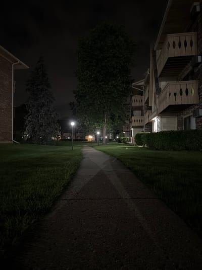 A paved walkway curves through a grassy area at night, illuminated by streetlights. To the right, a multi-story brick apartment building with balconies is visible. To the left, another brick building and several trees are present. The sky is dark and overcast. The location is the city of Vernon Hills, United States of America.