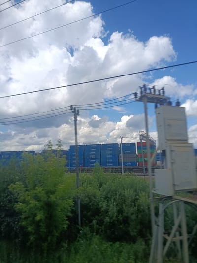 The image shows a view of a railway yard in Березка-291 Снт, Russia, with numerous shipping containers stacked high. The containers are predominantly blue, with some green and red ones visible. Some containers have logos and text, including "PANDA EXPRESS LINE" and "MAXHUN." Overhead power lines traverse the sky, and a utility pole with electrical equipment stands in the foreground, out of focus. Lush green foliage and trees line the foreground and middle ground, obscuring the lower parts of the containers and railway tracks. The sky is partly cloudy, with bright blue patches and large, fluffy white clouds. The overall impression is one of an industrial area on a bright, clear day. There are no people visible in the frame.