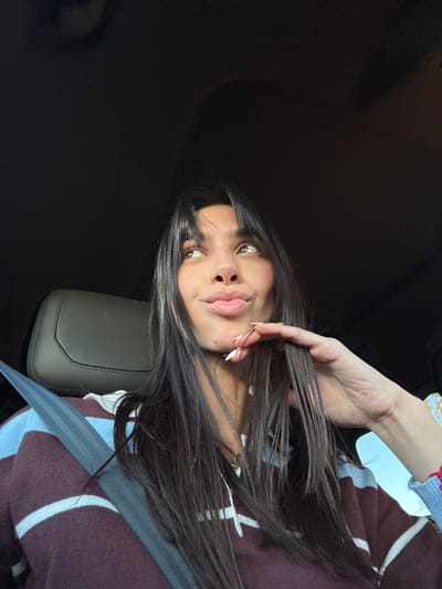 A young woman with long, dark hair and light skin is looking into the camera. She has a neutral expression. Her left hand is raised, and she is touching her hair. She appears to be sitting in a car, wearing a sweater with blue and white stripes. A seatbelt is visible across her torso. The background is dark, suggesting the car's interior.