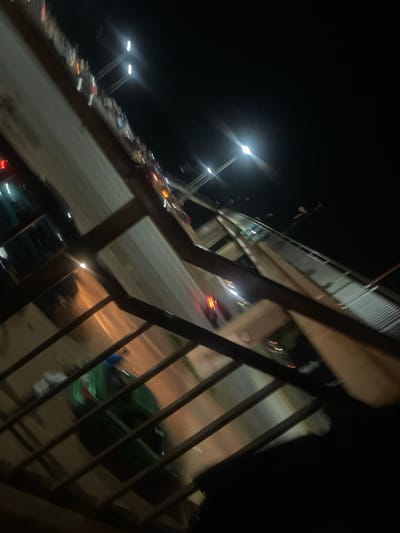 The media file shows a nighttime scene with a road and vehicles, likely in Sauka, Nigeria. The perspective is from an elevated vantage point, possibly a pedestrian bridge or balcony, looking down onto a street where cars are moving. The lighting is predominantly from streetlights and the headlights and taillights of the vehicles, creating streaks of light due to the motion and possibly a long exposure.
Several streetlights are visible, some appearing as bright, elongated glows. The road is paved, and the surrounding area is dark, indicating it's nighttime. The blurriness of the image suggests movement, either of the camera or the vehicles, or both.
On the road, there are multiple vehicles, appearing as indistinct shapes with illuminated lights. Some red taillights are visible, indicating cars moving away, and white headlights suggest cars approaching. One larger, dark green vehicle is somewhat discernible through a metal railing in the foreground.
In the foreground, dark metal railings create a visual barrier, framing the scene below. The overall impression is that of an urban or suburban environment at night, with traffic flow. The time of day is clearly night, and there are no obvious cues about the weather or specific emotions conveyed. No visible text is present in the image. The scene depicts the general activity of traffic on a road at night.