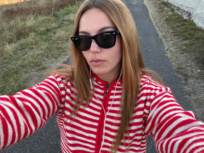 A person is wearing a red and white striped jacket and black sunglasses, with light brown hair and headphones. The picture appears to be taken at an upside down angle, showcasing the person's face, upper torso and surrounding environment. The background shows asphalt and grassy terrain, suggesting an outdoor setting. The lighting is natural, indicating it is daytime.