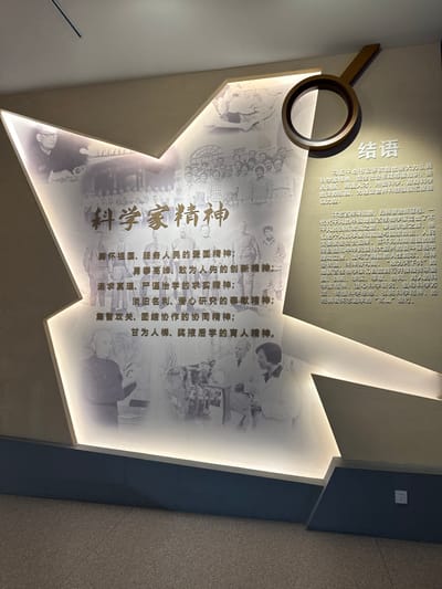 The image depicts a wall display featuring the "Spirit of Scientists." It includes Chinese text about patriotism, innovation, truth-seeking, detachment from fame, teamwork, and mentorship. There is a magnifying glass as an accent. Additional text mentions President Xi Jinping's call to promote the spirit of scientists and foster a nation dedicated to science and technology. The display shows historical black and white images of people working in a scientific setting.