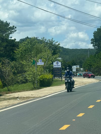 The image shows a road in Thôn 6, Vietnam, with a motorcyclist riding on the right side of the frame. The road is paved with asphalt and has yellow lane markings. On the left side of the road, there is a sign for "DOIDEP" which appears to be a resort and spa, restaurant, tea & coffee shop, and showroom. There are also trees and foliage on either side of the road. In the background, there are hills covered with trees. The sky is cloudy. The motorcyclist is wearing a helmet and a mask, and is riding a black motorcycle. There are other vehicles in the distance, including a red car and a few other motorcycles. The scene appears to be captured during the daytime, with moderate lighting.