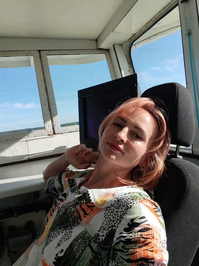 A woman with reddish-pink hair is seated in what appears to be a control room or cabin, likely on a vessel or in a tower, given the panoramic windows. She is looking towards the camera with a relaxed expression, her head tilted slightly and her left hand resting near her chin. She is wearing a colorful, patterned shirt with a mix of abstract shapes and animal prints in green, orange, white, and black.
The setting features large windows with visible metal frames, suggesting a robust structure designed for a wide field of view. Outside the windows, a bright blue sky with sparse clouds indicates pleasant weather during the daytime. A distant, tree-lined shore can be seen, hinting at a location near a body of water or a vast landscape. Inside, a dark monitor is positioned behind her, and a black headrest is visible to her right. Some utilitarian equipment or controls are partially visible in the lower left of the image.
The overall mood is calm and contemplative, with the sunlight creating highlights on the woman's face and hair. The presence of the windows and the view of the natural landscape suggest the scene is set in a functional location with a connection to the outdoors. No visible text is apparent in the image.