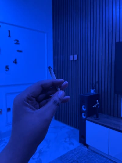 The image, taken in Alozo, Nigeria, captures a close-up of a human hand holding a lit, hand-rolled cigarette or joint, with ash visible at its tip. The scene is illuminated by a strong blue light, casting the entire indoor environment in a monochromatic hue, suggesting nighttime or a dark room with artificial lighting.

The hand, with visible knuckles and fingers, holds the item steadily. In the background, a slatted wall or ceiling runs horizontally, providing a textured element to the scene. Above and to the right of the hand, a black electronic device, possibly a speaker or part of a sound system, is visible with circular indentations or buttons on its face. Further in the background, a white surface displays distinct black numbers "1, 2, 4, 5," indicative of a clock face or a numbered game board. The subdued blue lighting creates an intimate and perhaps relaxed atmosphere for the individual smoking.