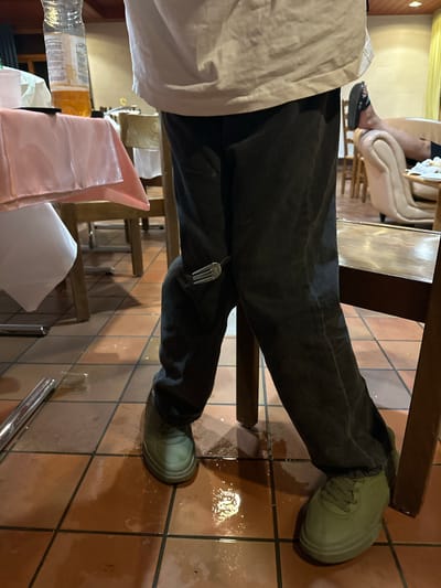 The image shows a person's lower body in a dimly lit indoor setting. The person is wearing dark pants and light green sneakers. A fork is stuck in the front of the right pant leg. The floor is tiled with brown squares and appears wet. There are parts of tables covered with pink and white tablecloths, wooden chairs, and furniture in the background, suggesting a restaurant or dining area. A plastic bottle with yellow liquid is visible on a table. The scene does not provide any specific visual cues to indicate the location as Wald (ZH), Switzerland.