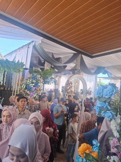 The image depicts a scene in an outdoor gathering space in Bojongsoang, Indonesia (latitude: -6.968, longitude: 107.651). The space is covered by a white and grey canopy, with a wooden ceiling above it. A large group of people, mostly women and children, are standing in a line. Several men stand behind them. The people are wearing clothing typical of the region. There are also various floral decorations in the scene, including bouquets of blue and pink flowers, as well as an elaborate arrangement with yellow, orange, and blue flowers. Some people are holding cups in their hands. There are also a few other objects in the scene, including a large mirror, a small table, and a few chairs. The scene appears to be a social gathering, perhaps a wedding reception or a celebration. The people appear to be interacting with each other in a friendly manner.