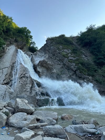 The image captures the Khuchni Waterfall in Khuchni, Russia, under a clear blue sky. A powerful waterfall cascades down a rugged, rocky cliff face. The water is white and turbulent, crashing into a pool at the base where it churns into a foamy spray. Large boulders are scattered around the pool, some partially submerged. The surrounding landscape is steep and covered in lush green trees and vegetation. The rocky slopes are also dotted with plants. In the foreground, closer to the viewer, are more large rocks and debris, including plastic bottles and other litter scattered on the ground. The scene depicts a natural, wild environment with signs of human impact from the trash.