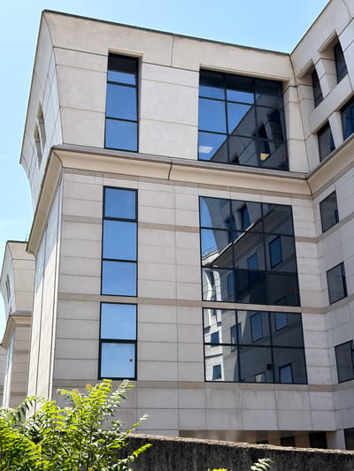 This is an eye-level, slightly angled, close-up, outdoor photograph of a modern building with a light-colored stone facade and numerous dark-tinted windows. The building appears to be an office building or a similar commercial structure. The windows are arranged in a grid pattern, with some reflecting the sky and other parts of the building. Some windows are larger and rectangular, while others are smaller and square. There are some geometric architectural elements protruding from the building's surface.
In the lower right corner, a portion of a tree with vibrant green foliage is visible, suggesting it is daytime and likely spring or summer. The sky is a clear, bright blue, indicating fair weather. The time of day appears to be mid-morning or early afternoon, given the lighting and shadows. There is no text visible in the image. The scene evokes a sense of urban modernity and architectural design.