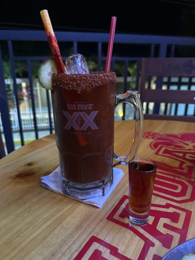 A large mug of Dos Equis beer sits on a wooden table, rimmed with chili salt, garnished with a lime and tamarind candy stick, and two red straws. A shot glass with a brown liquid sits beside it on a white napkin. Red branding is visible on the table. A metallic railing and buildings are visible through a window in the background.