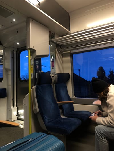 Inside a train, a seated person uses a phone. Two rows of blue patterned seats sit adjacent to windows showing a blue sky with silhouetted trees and structures. Overhead are air conditioning vents. A suitcase rests near the seats. The train interior is primarily white, with some yellow and metal accents. Another window is visible toward the end of the train car.