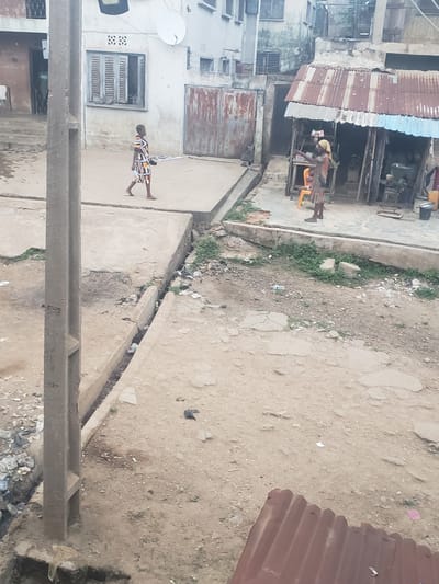 The image captures a street scene in Alalubosa, Nigeria. A dirt road is visible, with scattered debris and vegetation. On the left, there are buildings with corrugated metal roofs. One building appears to be a shop with a man standing inside. A person in a patterned dress is walking in the opposite direction on a paved area. A concrete structure runs along the bottom of the image, possibly a utility pole support.