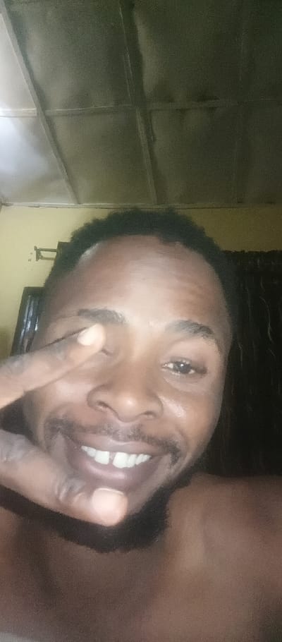 A man with dark skin, a full beard, and short black hair smiles with his finger slightly covering one eye. His teeth are visible, showing a small gap in the front. He appears to be shirtless. The background includes a textured ceiling, a light-colored wall, and a dark curtain or backdrop. Lighting is slightly dim, creating a shadowed effect on the man's face and surroundings.