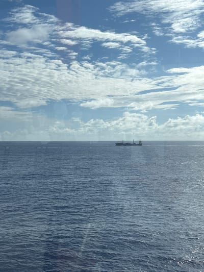 The image displays an expansive view of the open ocean and sky, observed through a reflective pane, likely a window, from the city of Long Island, Bahamas. The lower two-thirds of the frame consists of deep blue, mildly rippled ocean water with intermittent sunlight reflections. A dark-hulled cargo ship, featuring discernible superstructure and deck containers, is positioned on the distant horizon, slightly right of center, in transit across the water. The upper third of the image shows a blue sky populated by numerous white cumulus and stratocumulus clouds. No individuals are present in the scene.