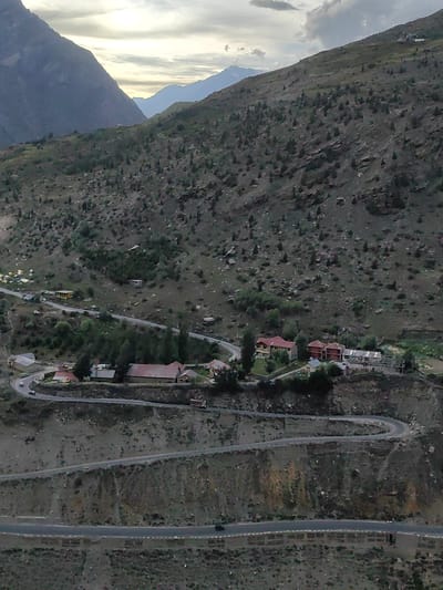 The image shows a winding road on a steep, rocky mountainside in Gushal, India. The road has multiple switchbacks and appears to be a paved surface. Several buildings, including houses and what appear to be commercial structures, are visible nestled on the slopes and at the base of the mountain. Some vehicles, including cars and a truck, are on the road. The environment is mountainous and arid, with sparse vegetation consisting of small trees and shrubs scattered across the rocky terrain. The sky is overcast with some clouds visible, and the lighting suggests it is either early morning or late afternoon. The overall scene depicts a remote, mountainous settlement with a challenging road infrastructure.