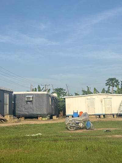 The image shows a scene in Igwuruta, Nigeria. The environment consists of buildings, open grassy terrain, and a blue sky with sparse clouds. Some buildings appear to be temporary or prefabricated structures; one is painted white while others are grey. Electrical wires are visible in the upper-left quadrant of the image, indicating an urban setting. A parked motorcycle sits near the white building. Some chairs and other items are partially covered next to the motorcycle. The grass appears to be wild and recently rained on.