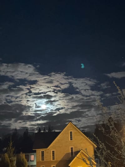 The image appears to be a night scene, possibly taken in or near a residential area. The sky is predominantly dark, with patches of clouds that reflect light, presumably from the moon, which is a bright spot visible through the clouds. There is a bright, teal object in the dark sky, presumably some kind of lit object. A building is partially visible on the right side, with several windows. Branches from a tree are visible on the top right of the image. The scene appears serene and quiet, likely taken at night in a residential area. The exact location in Лесной Пейзаж Кп (шарапово, Russia) cannot be determined from the image alone.