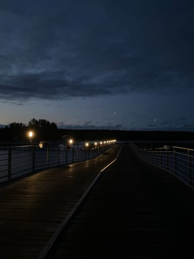 The image depicts a wooden pier at night in Kirkkolakhti, Russia. The pier is illuminated by a series of lights, casting a warm glow on the wooden planks. The sky is dark and cloudy, with a hint of twilight remaining. On the left side of the pier, a railing is visible, and beyond it, a body of water. In the distance, a line of trees marks the shoreline. The pier extends into the distance, disappearing into the darkness. The scene is tranquil and serene, with a sense of solitude.